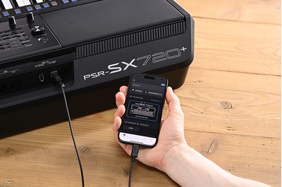 Yamaha Workstation Arranger PSR-SX720+ e una mano che tiene uno smartphone con la schermata dell’app ufficiale Yamaha “Expansion Explorer”.