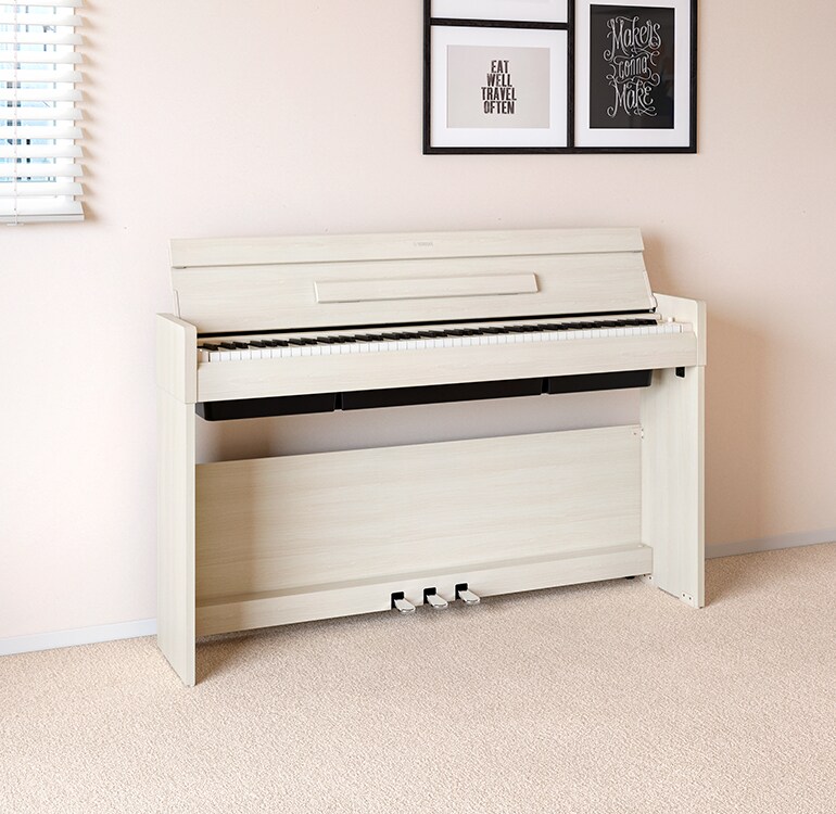 Un soggiorno con un pianoforte ARIUS in betulla bianca posizionato accanto a mensole a muro su cui si trovano lampade e un diffusore, armoniosamente integrato con l’arredamento.